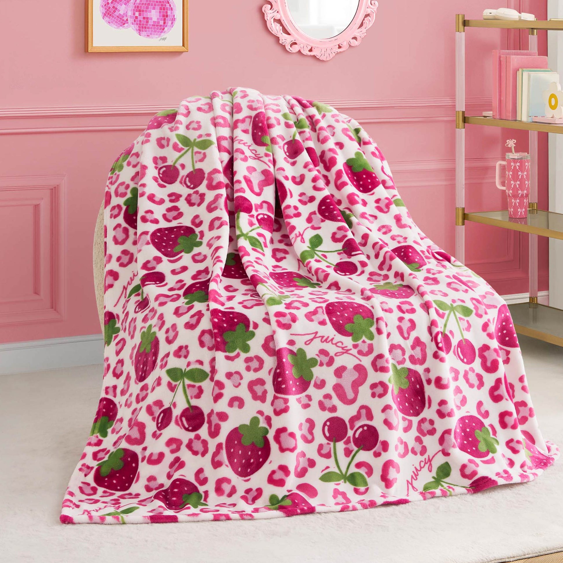 A Kids Plush Throw Blanket with pink leopard spots, strawberries, cherries, and green leaves is draped over a chair in a pink room with gold shelves and framed artwork on the wall.