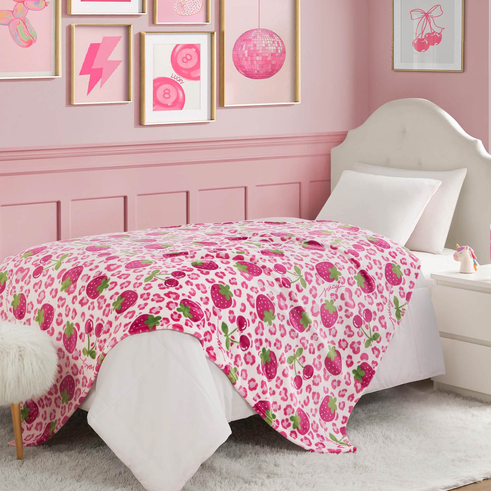 A twin bed with a white headboard and bedding showcases a Kids Plush Throw Blanket in pink and white strawberry and leopard print. The pink-themed room also features framed wall art and a small white nightstand.