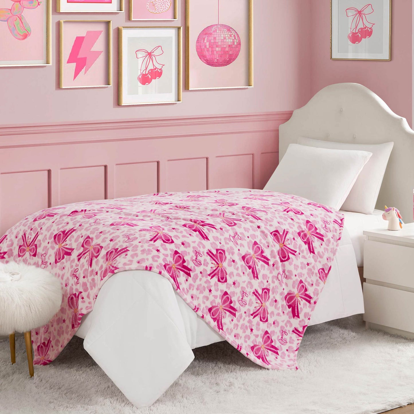 A neatly made bed with a Kids Plush Throw Blanket featuring ballet slipper designs brightens this pink-themed bedroom, paired with framed wall art, a fur-topped stool, and a nightstand holding a book and lamp.