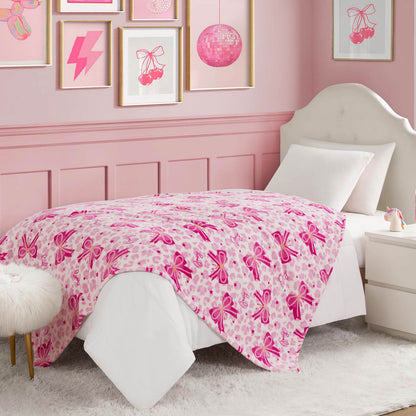 A neatly made bed with a Kids Plush Throw Blanket featuring ballet slipper designs brightens this pink-themed bedroom, paired with framed wall art, a fur-topped stool, and a nightstand holding a book and lamp.