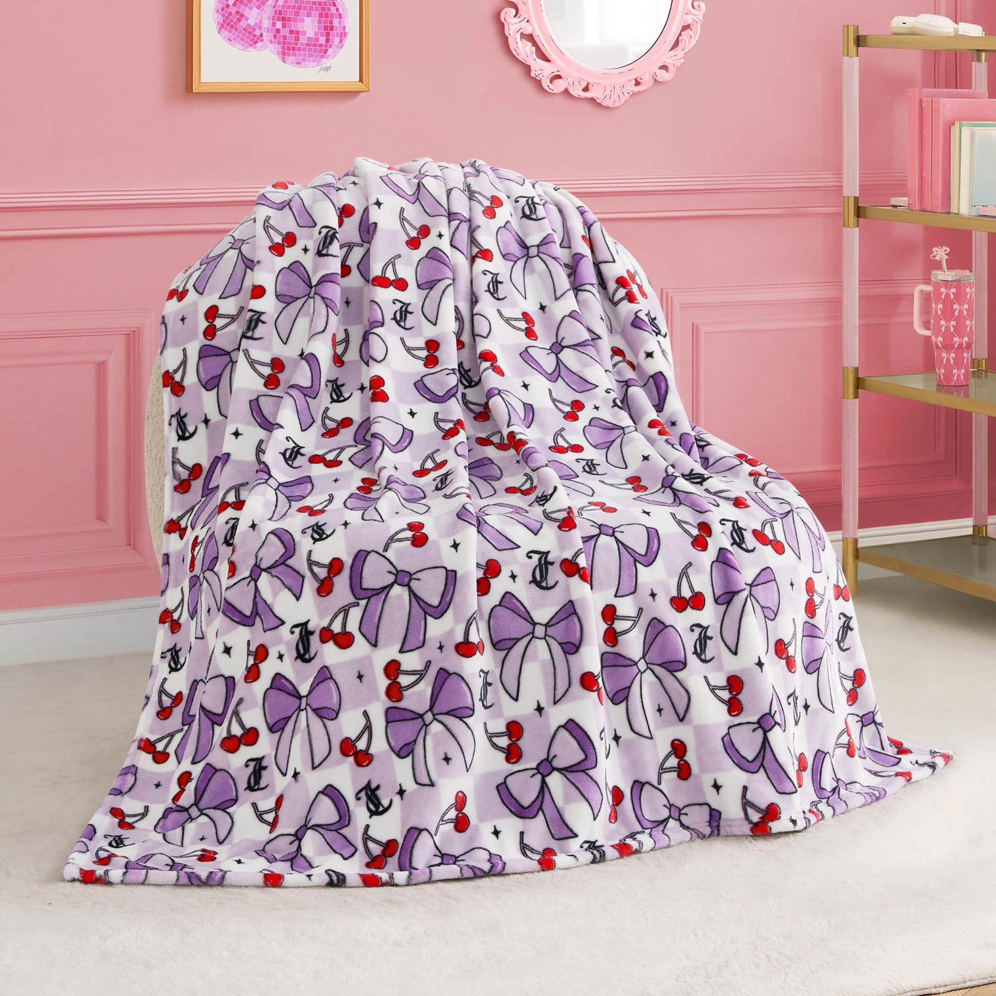 A Kids Plush Throw Blanket featuring purple bows, red cherries, and black anchors is draped over a chair in a pink room decorated with elegant accents and wall art.