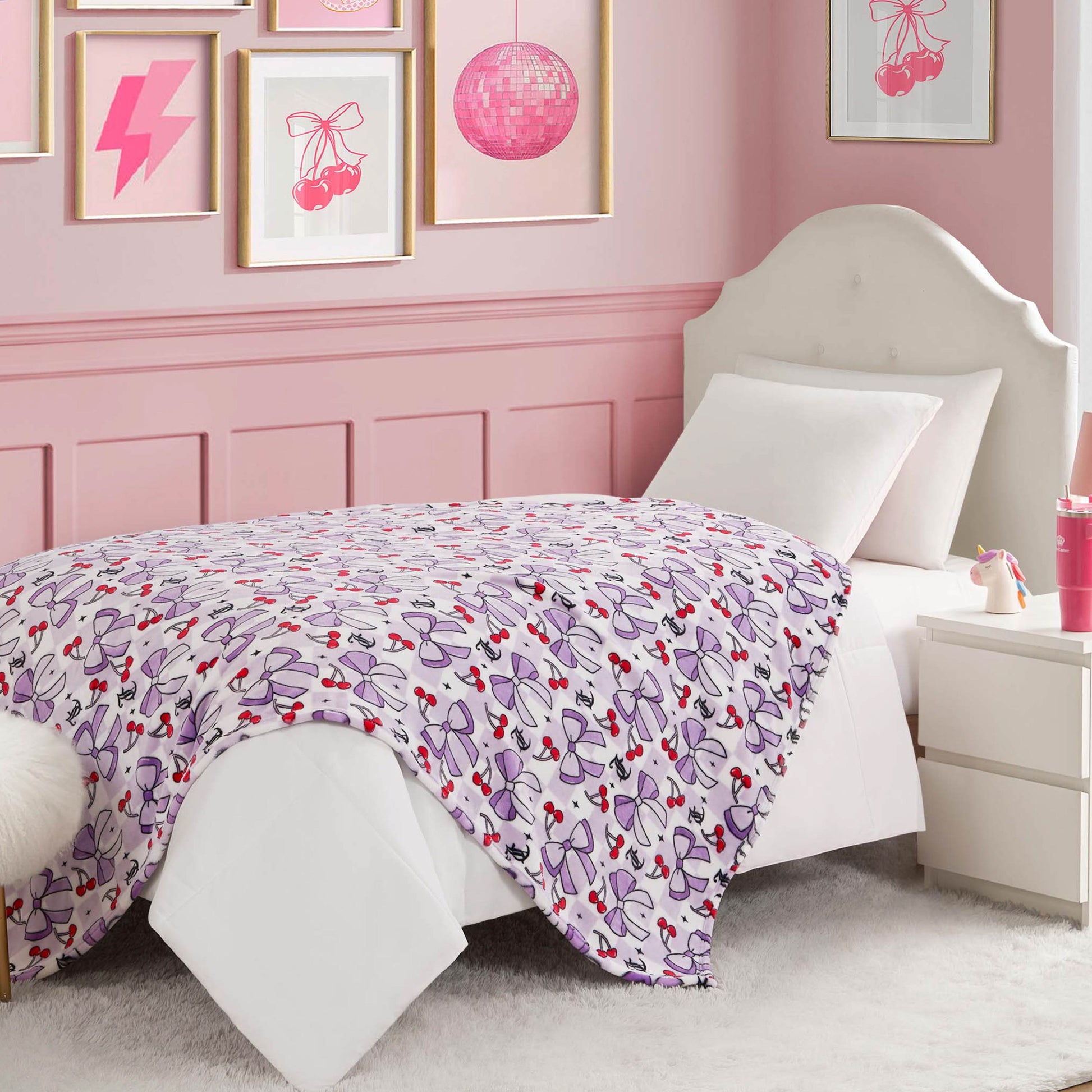 A neatly made bed with a white headboard features a Kids Plush Throw Blanket with purple and pink bows, in a pink bedroom with framed wall art and a white nightstand holding a pink water bottle.