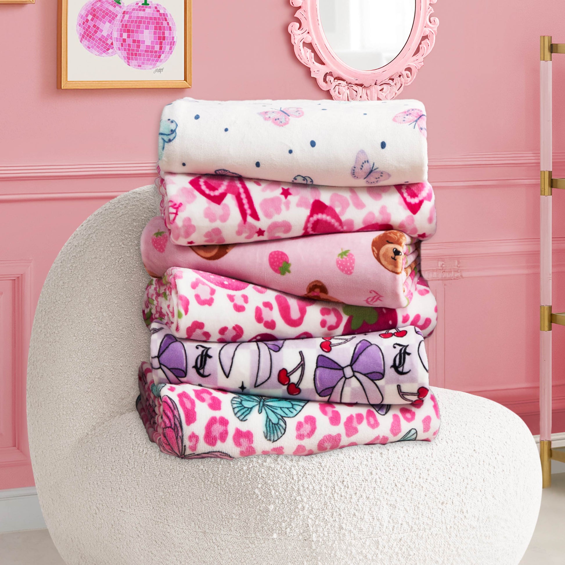 A stack of six Kids Plush Throw Blankets in colorful patterns is neatly folded on a textured white chair in a pink room with wall art, a round mirror, and gold accents.