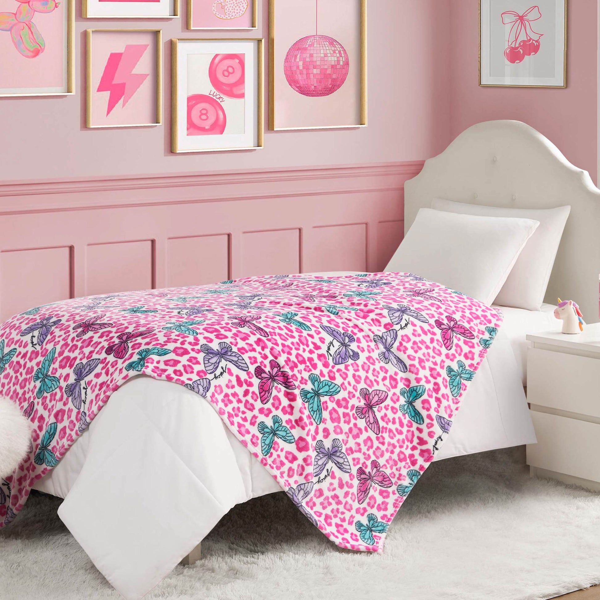 A pink-themed bedroom features a white bed topped with an ultra-soft Kids Plush Throw Blanket in pink leopard spots and blue bows. Pink art decorates the walls, and a white nightstand sits beside the bed on a fluffy rug.
