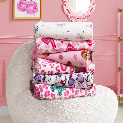 A stack of six Kids Plush Throw Blankets with fun designs sits on a textured white chair in a stylish pink room decorated with framed art and a wall mirror.