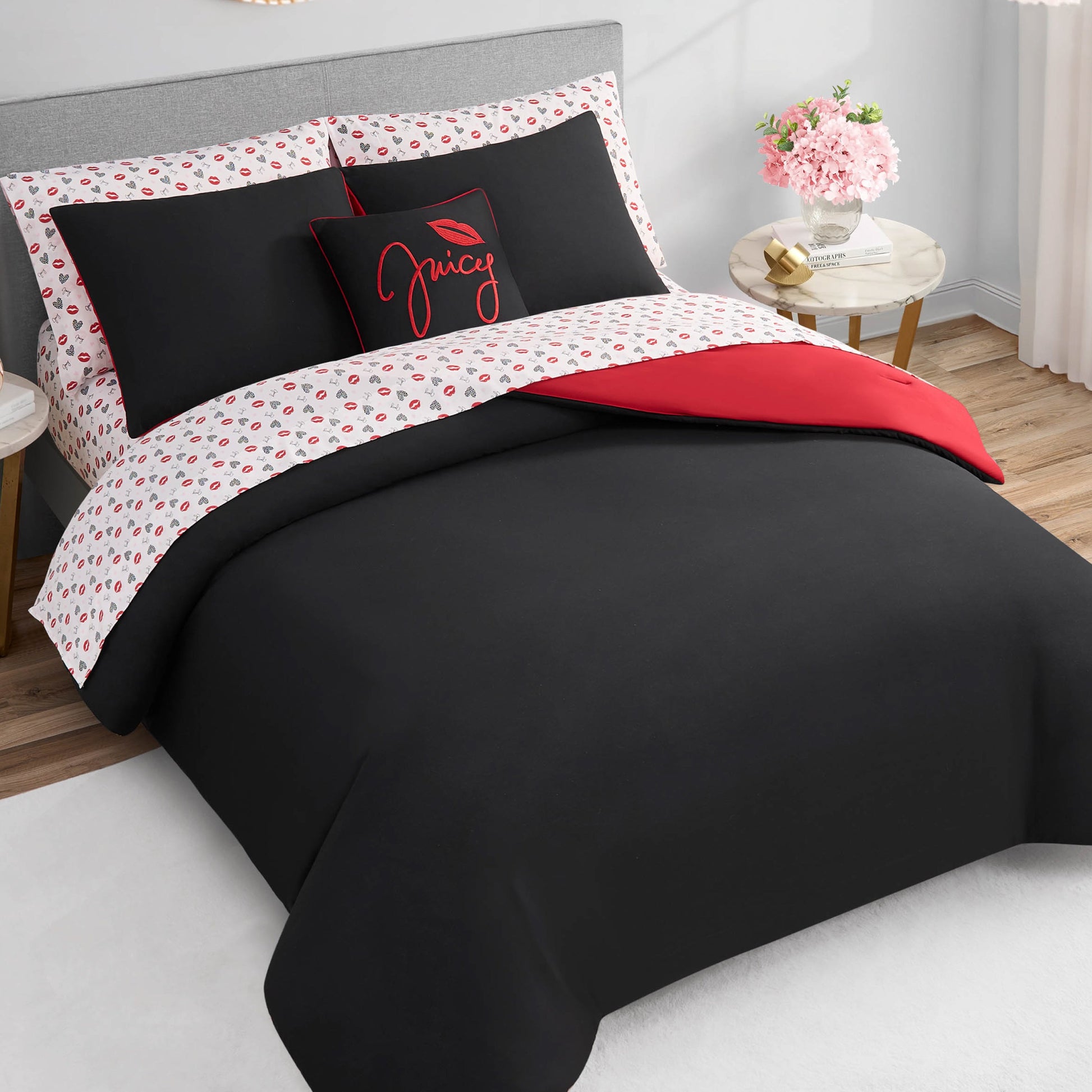 A neatly made bed features the Glam Girl Solid Reversible Bed in a Bag Bedding Set in black and red, a floral sheet, matching pillowcases, and a Juicy-accented decorative pillow. Next to the bed is a round table topped with pink flowers.