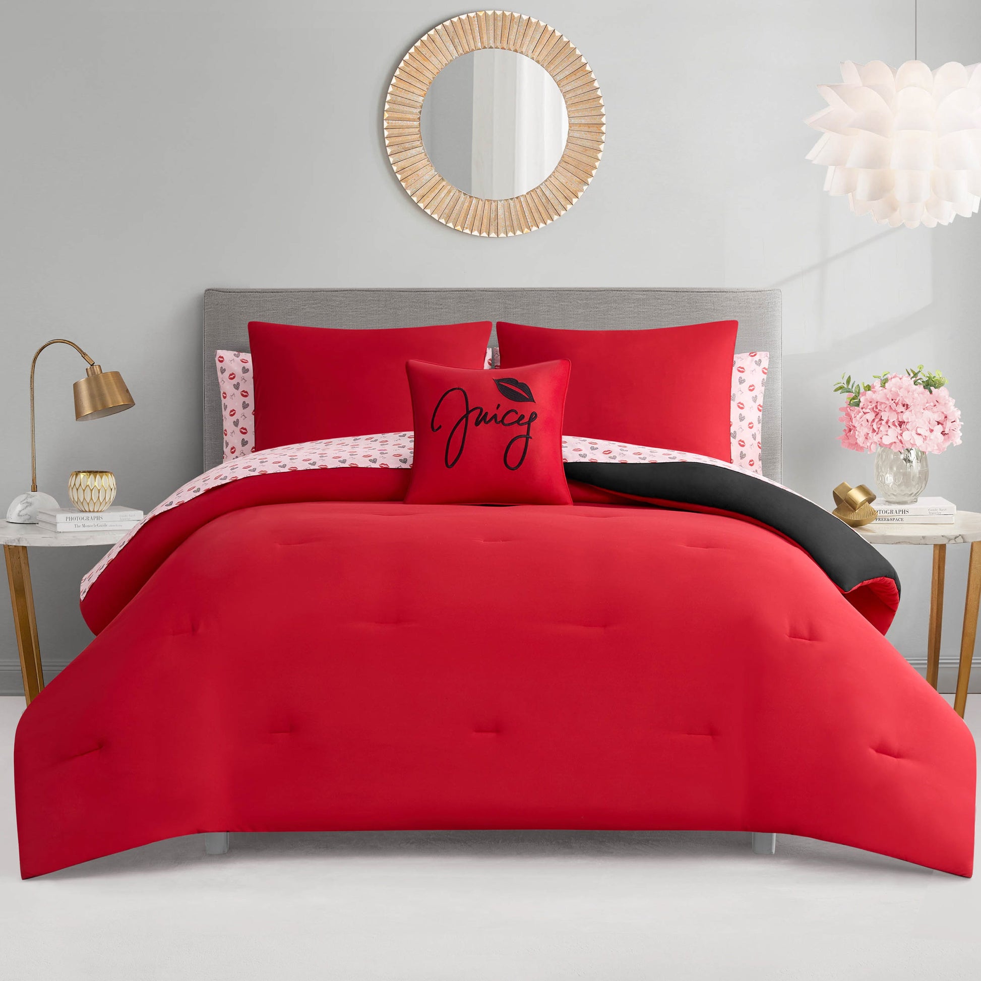 A modern bedroom featuring a gray upholstered bed, the Glam Girl Solid Reversible Bed in a Bag Bedding Set with bright red comforter, Juicy pillow, pink floral sheets, gold lamp, pink flowers, and a round mirror above the bed.