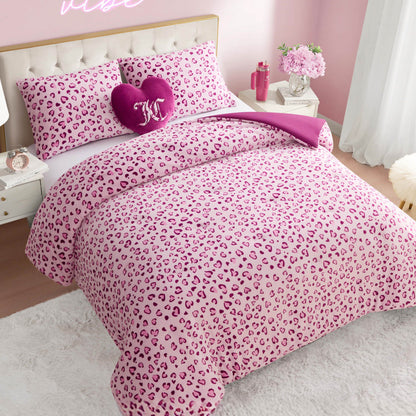 A bed styled with the Plush Reversible Leopard Hearts Comforter Set features a pink leopard print, matching pillows, a purple heart-shaped accent pillow with a K, white furniture, a fluffy rug, flowers, and sheer window curtains.