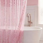 A white bathtub with a gold faucet sits behind a sheer pink PEVA Shower Curtain Liner in a softly lit bathroom with light pink walls and a window with white blinds.
