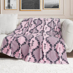 A Plush Oversized Throw Blanket by Juicy Couture, pink with a black crown and JC pattern, is draped over a white sofa. Ultra-soft and stylish, it adds flair to the room as framed photos hang on the wall behind.