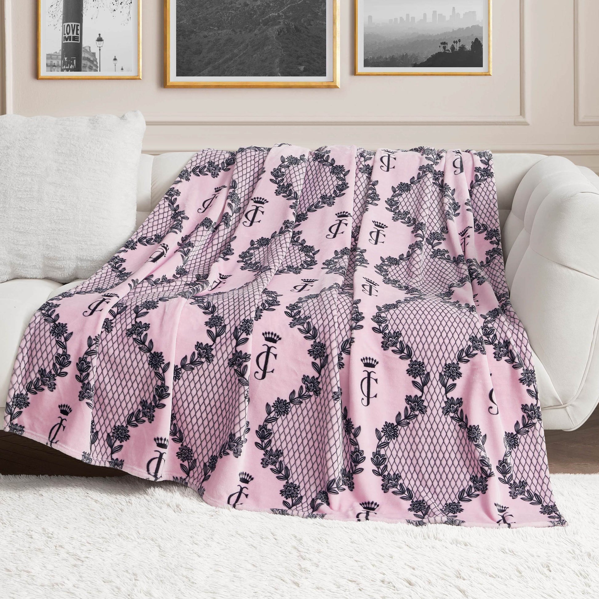 A Plush Oversized Throw Blanket by Juicy Couture, pink with a black crown and JC pattern, is draped over a white sofa. Ultra-soft and stylish, it adds flair to the room as framed photos hang on the wall behind.