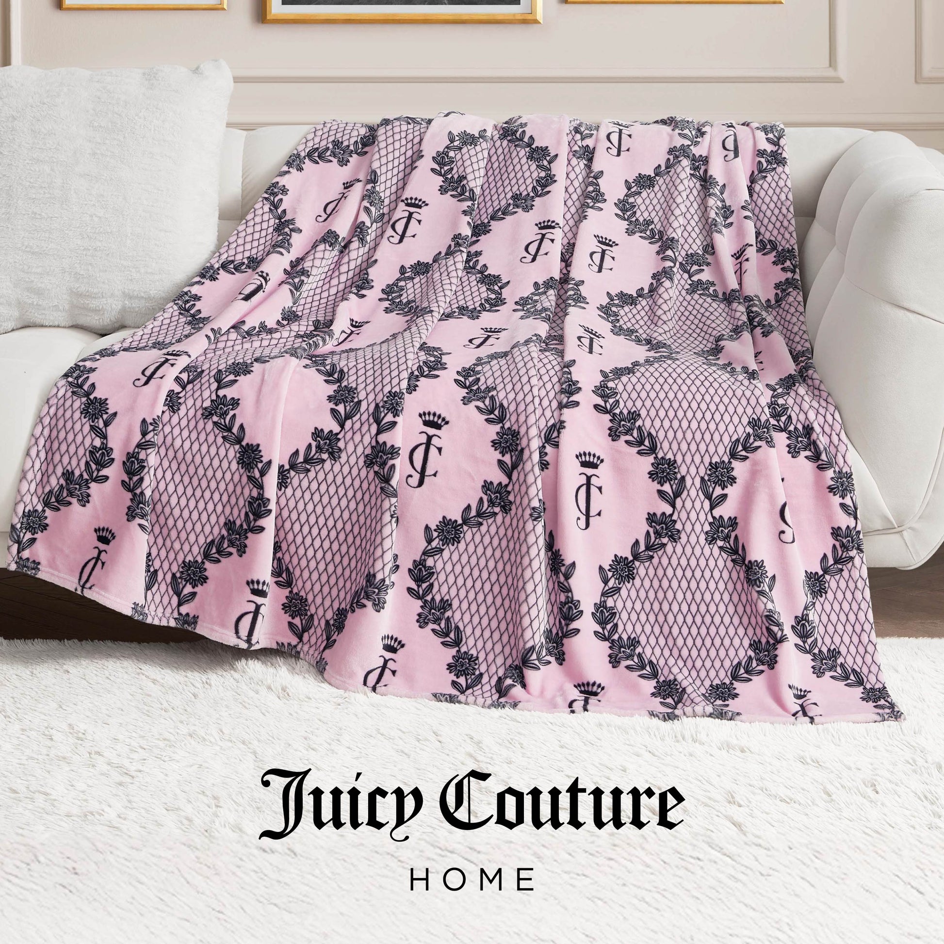 A pink Plush Oversized Throw Blanket with black crown and wreath patterns is draped over a white sofa. This ultra-soft blanket features "Juicy Couture HOME" at the bottom, adding a luxurious touch to the stylish living room.