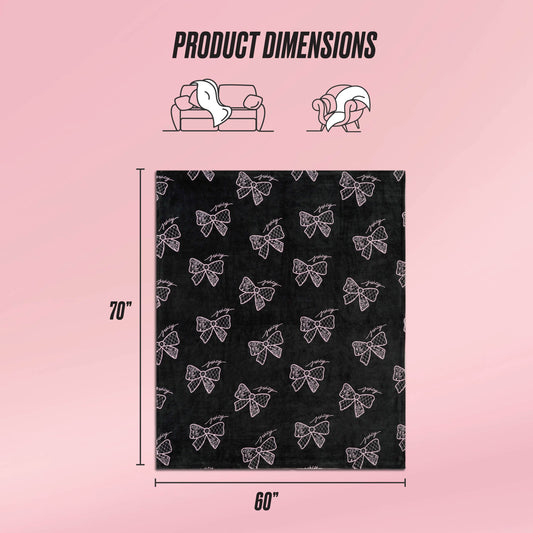 The Plush Oversized Throw Blanket by Juicy Couture is shown in black with a white bow pattern, measuring 70"x60". Ultra-soft and plush, it's ideal for couch or personal use, displayed against a pink background with helpful icons.