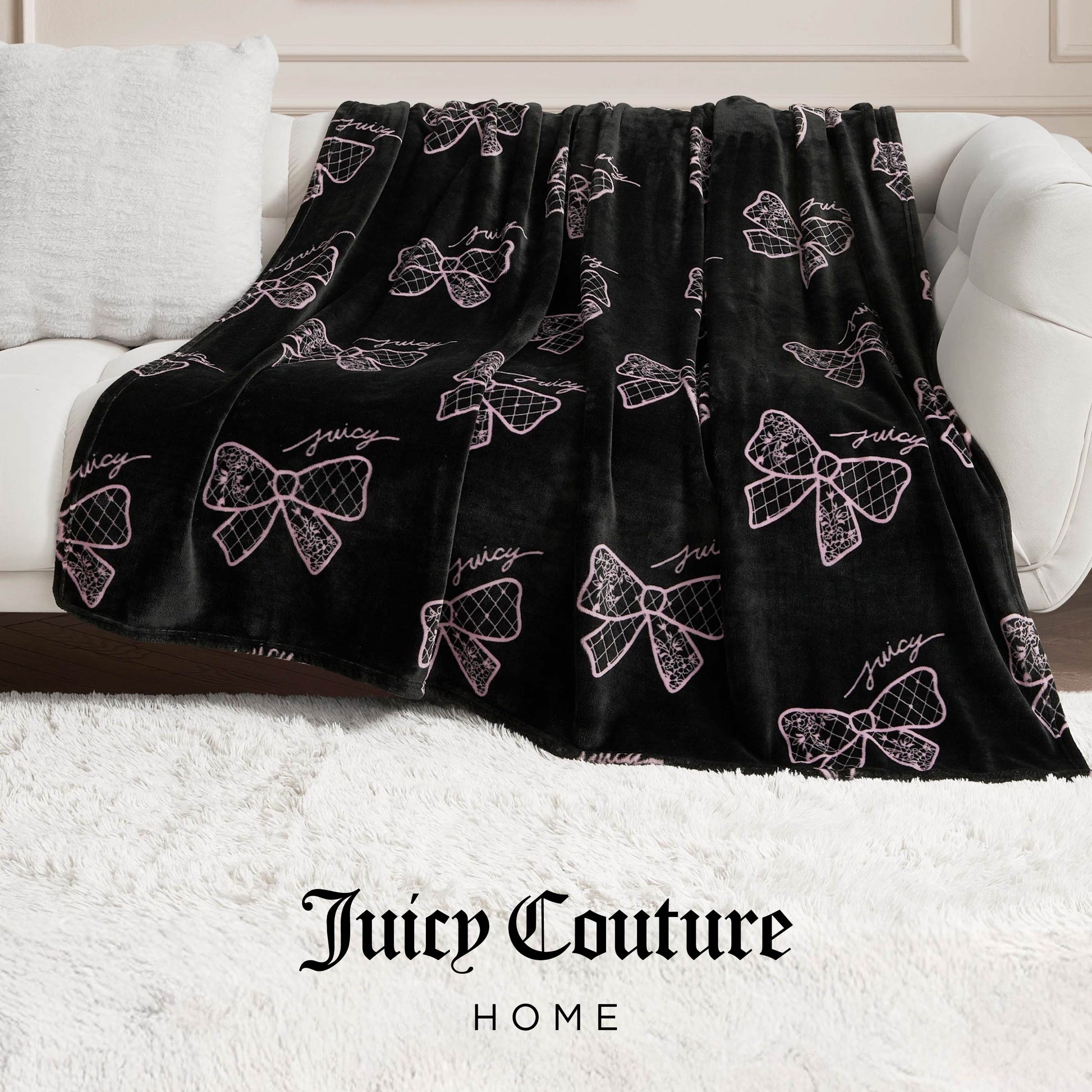 A black Plush Oversized Throw Blanket with pink bows and “Juicy” in cursive drapes over a white sofa on a fluffy rug, while “Juicy Couture Home” below spotlights this ultra-soft blanket’s cozy elegance.