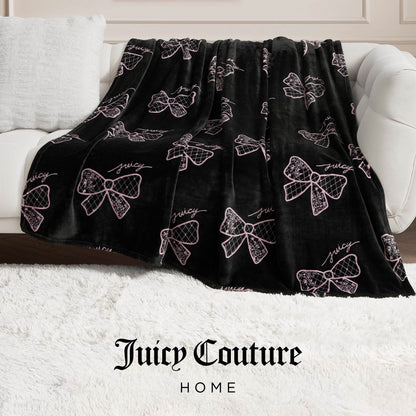 A black Plush Oversized Throw Blanket with pink bows and “Juicy” in cursive drapes over a white sofa on a fluffy rug, while “Juicy Couture Home” below spotlights this ultra-soft blanket’s cozy elegance.