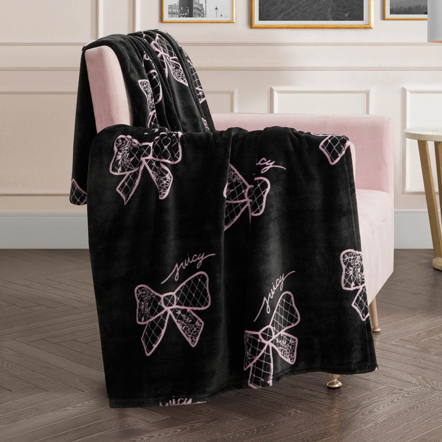 A Plush Oversized Throw Blanket, ultra-soft with pink bow designs and "Juicy" lettering, is draped over a pale pink upholstered chair in a modern room with light walls and framed pictures.