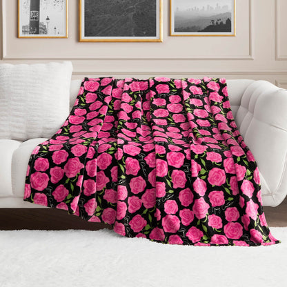 The Plush Oversized Throw Blanket, featuring vibrant pink roses and green leaves on a black background, is draped over a white couch in a bright, elegant living room decorated with framed artwork.