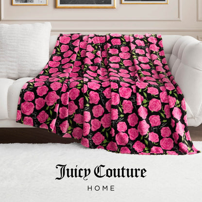 The Plush Oversized Throw Blanket, featuring a pink rose and green leaf design on black, is draped over a white sofa. "Juicy Couture Home" appears at the image's bottom.