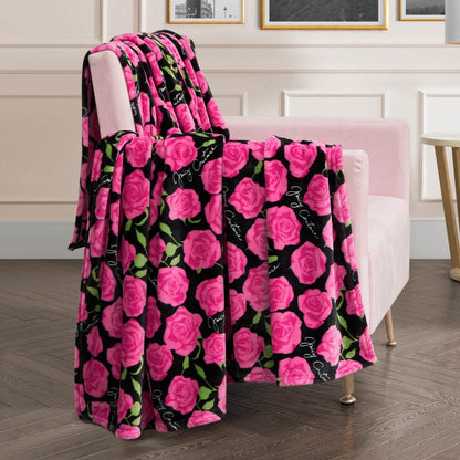 A plush pink chair features the Plush Oversized Throw Blanket, ultra-soft and decorated with vibrant pink roses, green leaves, and cursive white text on a black background in an elegantly lit room with framed artwork.