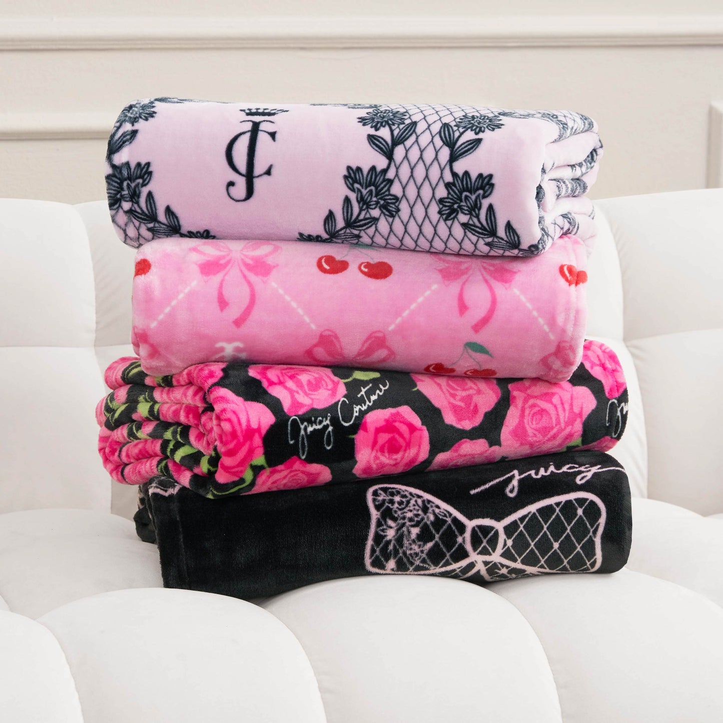 Four Plush Oversized Throw Blankets with pink and black patterns—cherries, roses, and “Juicy Couture” logos—are stacked on a white tufted cushion in a bright room.