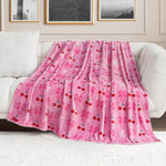 A Plush Oversized Throw Blanket by Juicy Couture, featuring pink fleece with red cherries, pink bows, and white monogram patterns, is draped over a white sofa in a modern living room with framed wall art.