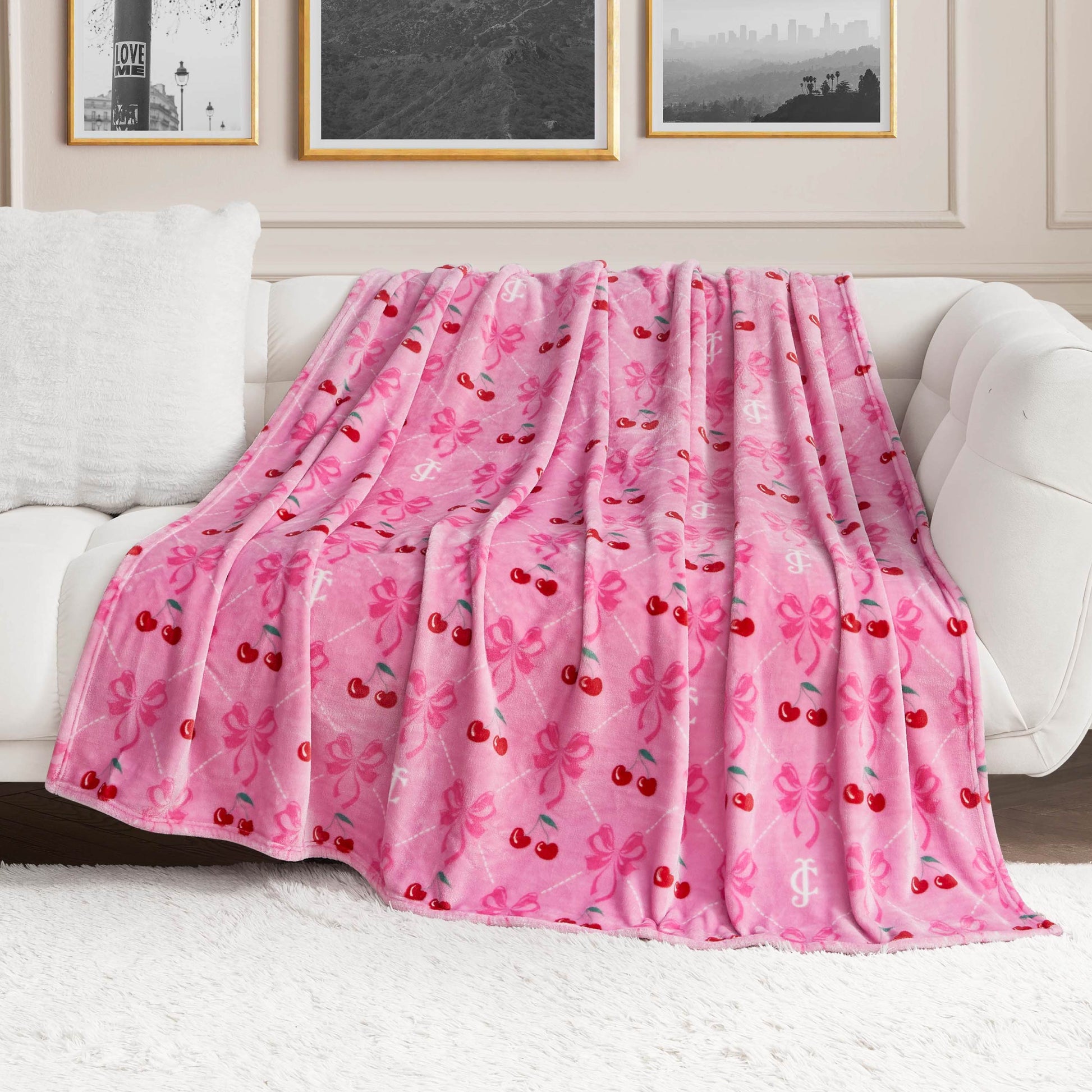 A Plush Oversized Throw Blanket by Juicy Couture, featuring pink fleece with red cherries, pink bows, and white monogram patterns, is draped over a white sofa in a modern living room with framed wall art.