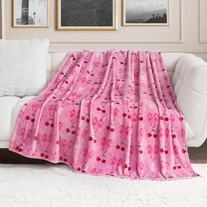 A Plush Oversized Throw Blanket by Juicy Couture, featuring pink fleece with red cherries, pink bows, and white monogram patterns, is draped over a white sofa in a modern living room with framed wall art.