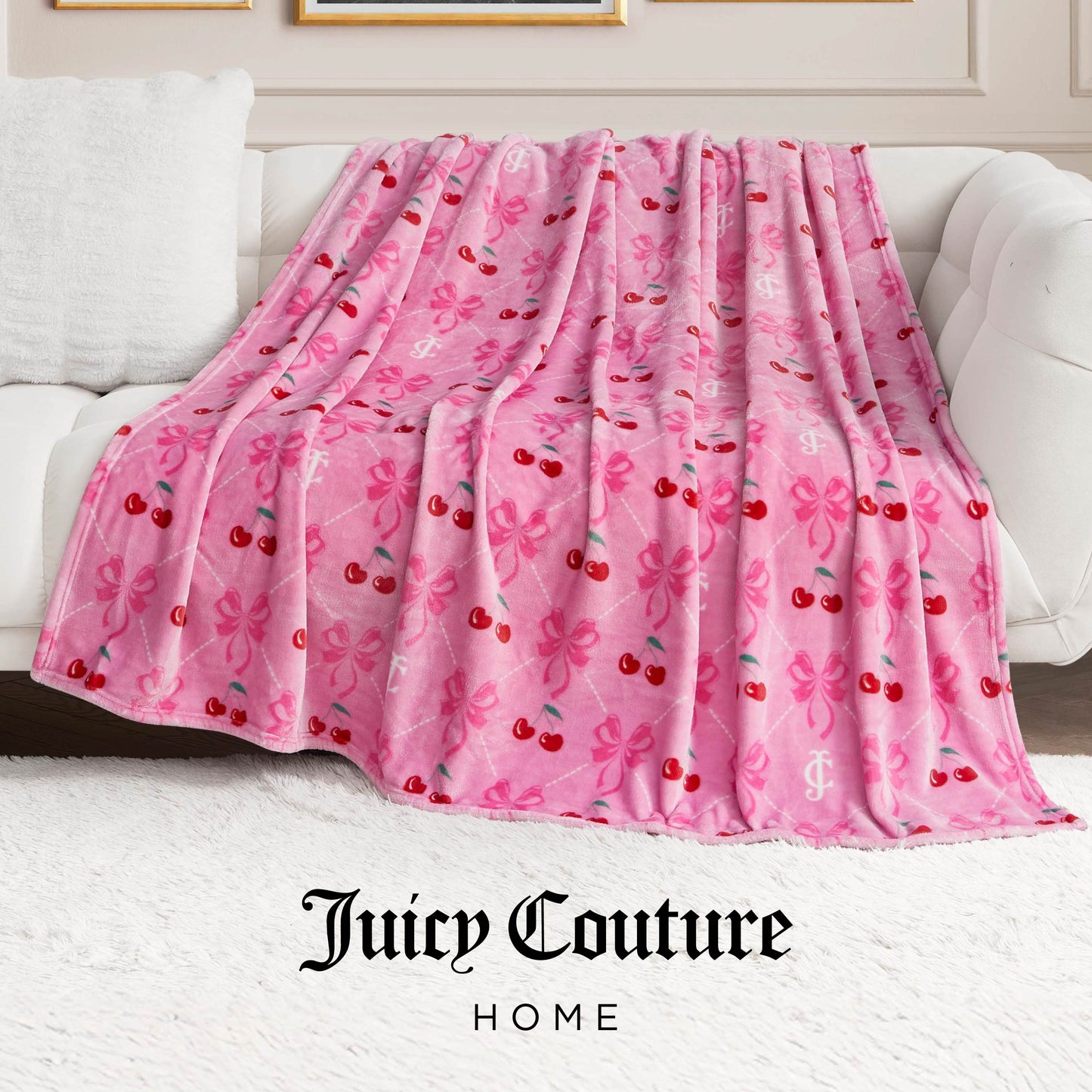 A Plush Oversized Throw Blanket with cherry and bow patterns is draped over a white sofa. The ultra-soft blanket displays the Juicy Couture Home logo at the bottom of the image.