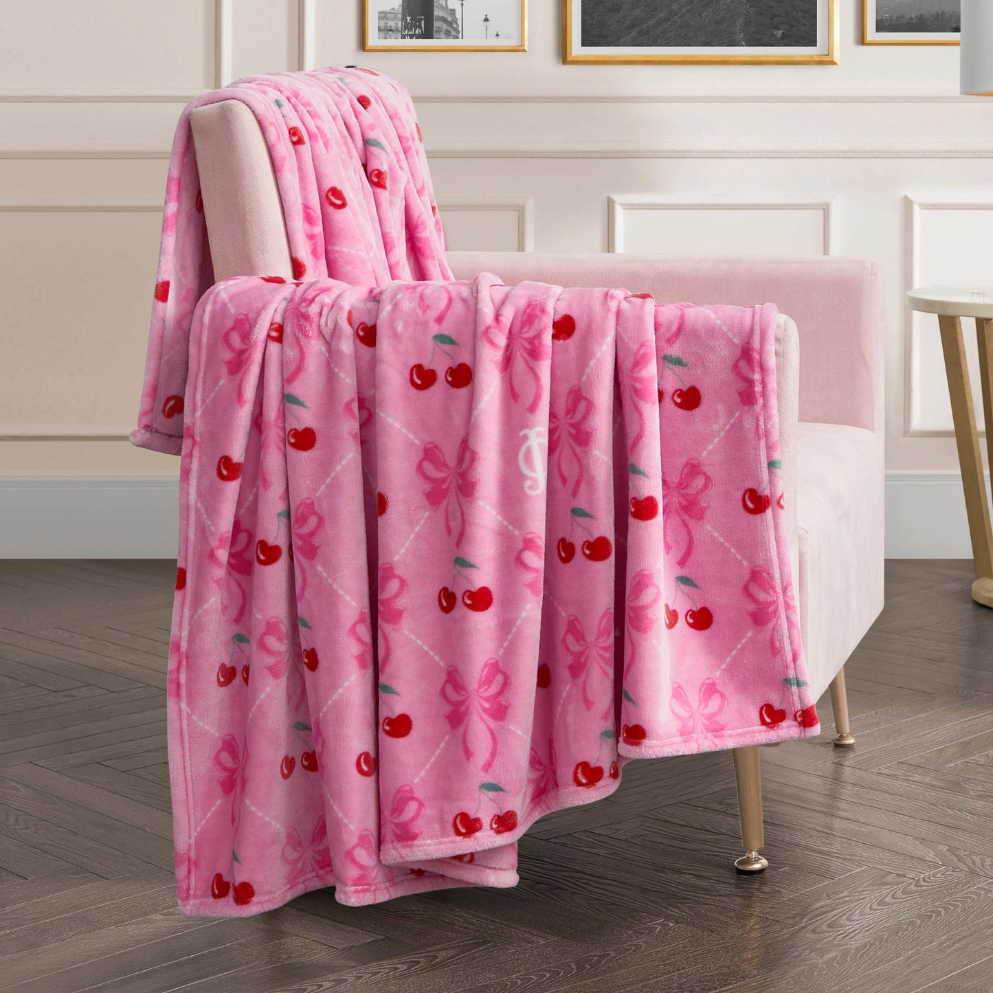 A pink armchair features the Plush Oversized Throw Blanket—ultra-soft with red cherries and pink bows—in a chic room with white paneled walls and framed art.