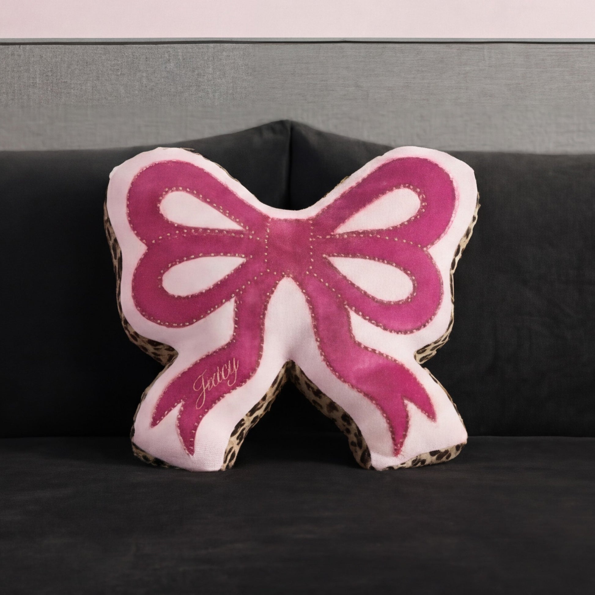 A Reversible Bow Decorative Pillow by Juicy Couture, pink with leopard print edges and "Juicy" embroidery, sits on a dark gray sofa against a matching gray wall.