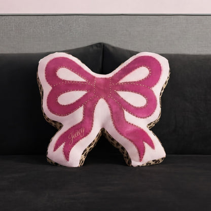 A Reversible Bow Decorative Pillow by Juicy Couture, pink with leopard print edges and "Juicy" embroidery, sits on a dark gray sofa against a matching gray wall.