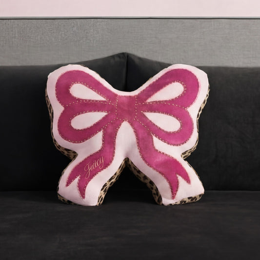 A Reversible Bow Decorative Pillow by Juicy Couture, pink with leopard print edges and "Juicy" embroidery, sits on a dark gray sofa against a matching gray wall.