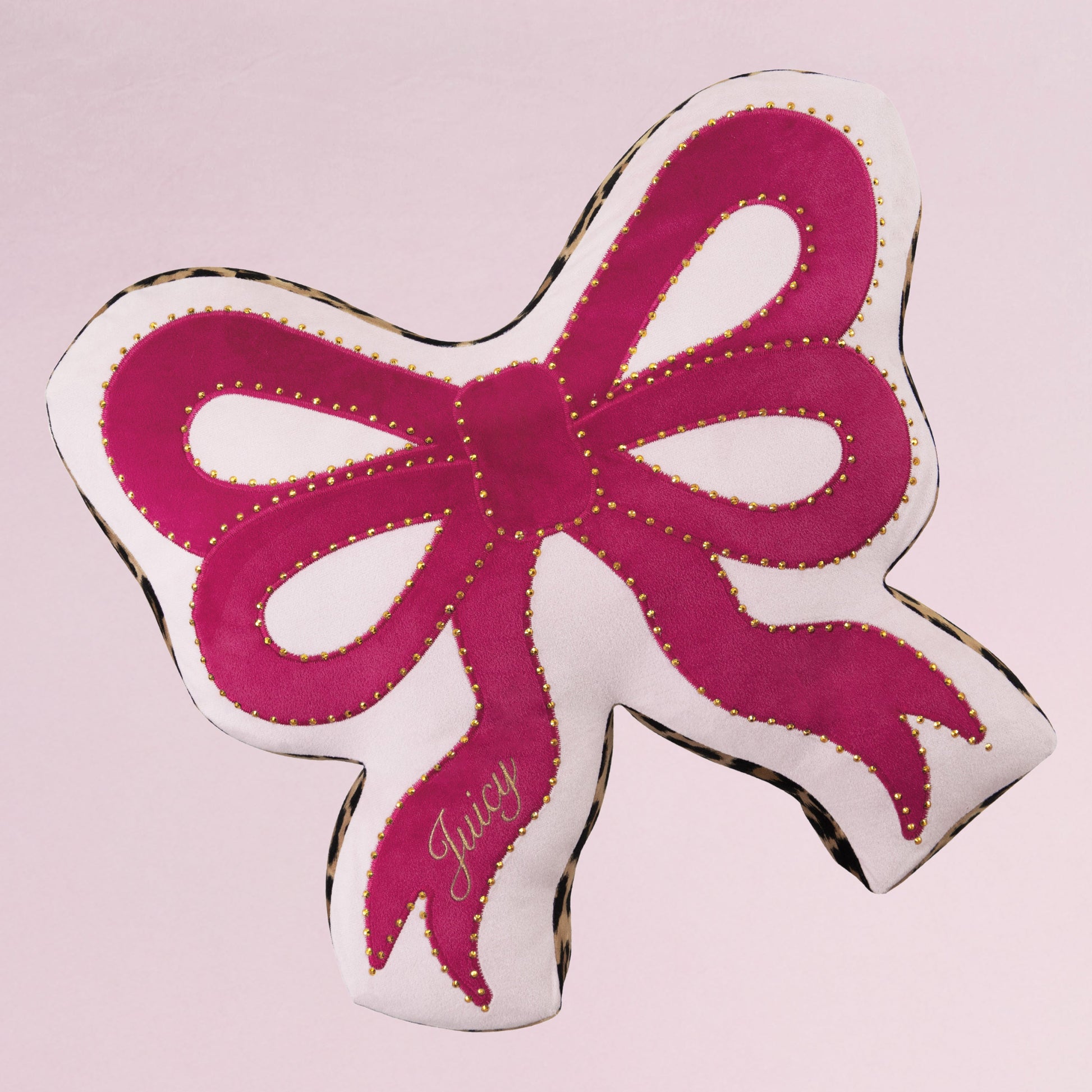 The Reversible Bow Decorative Pillow by Juicy Couture features pink and white fabric, gold stitching, and “Juicy” embroidered on one tail. Its bow shape adds a stylish touch to any space, set against a light pink background.