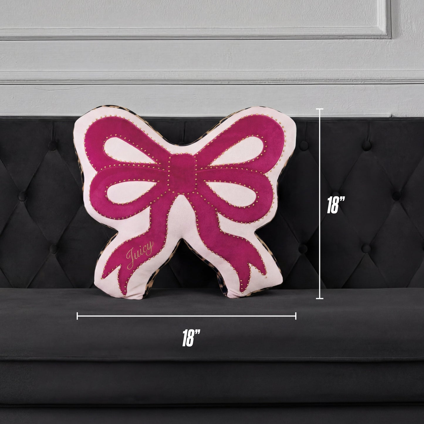 A Reversible Bow Decorative Pillow by Juicy Couture, pink and white with "Juicy" on it, sits on a black tufted sofa. Shaped like a bow, this decorative pillow measures 18" x 18".