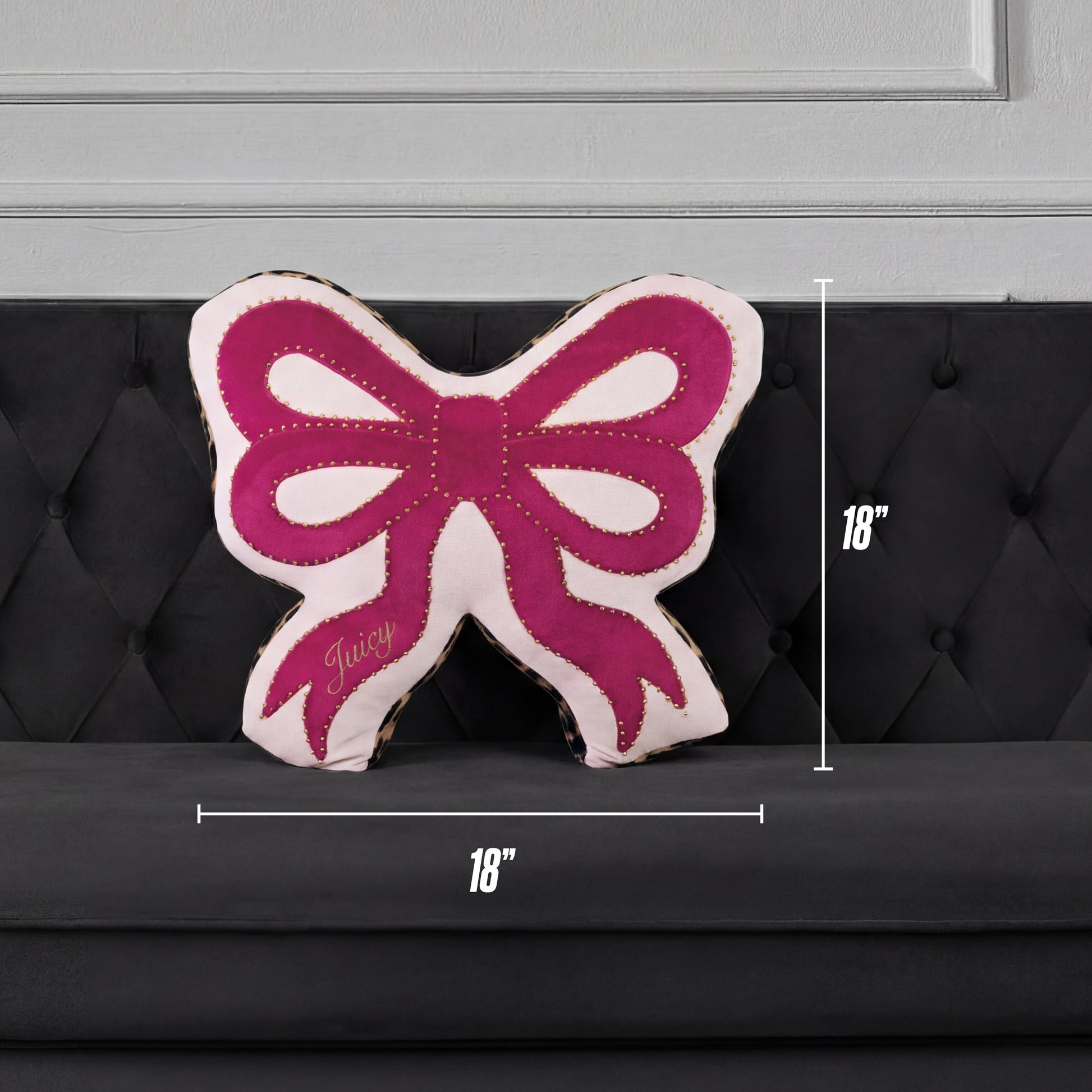A Reversible Bow Decorative Pillow by Juicy Couture, pink and white with "Juicy" on it, sits on a black tufted sofa. Shaped like a bow, this decorative pillow measures 18" x 18".