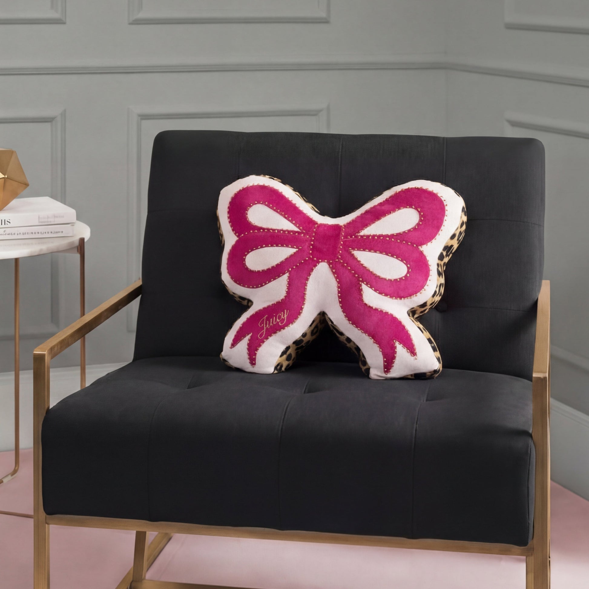A black chair with gold armrests holds the Reversible Bow Decorative Pillow by Juicy Couture, shaped like a pink and white bow with leopard print edges and "Juicy" embroidery. Light gray paneled walls form the background.
