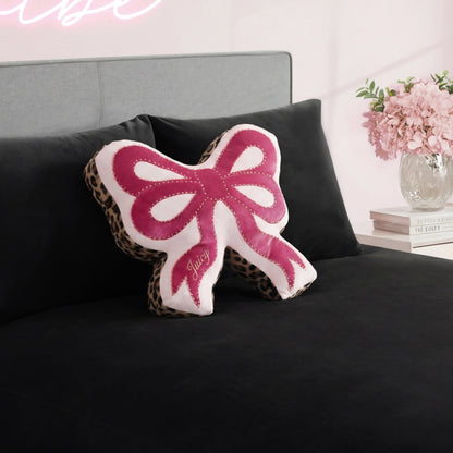 A Reversible Bow Decorative Pillow by Juicy Couture, featuring pink, white, and leopard print details, rests on a black bedspread. Pink flowers and books decorate the nearby nightstand in the softly lit bedroom.