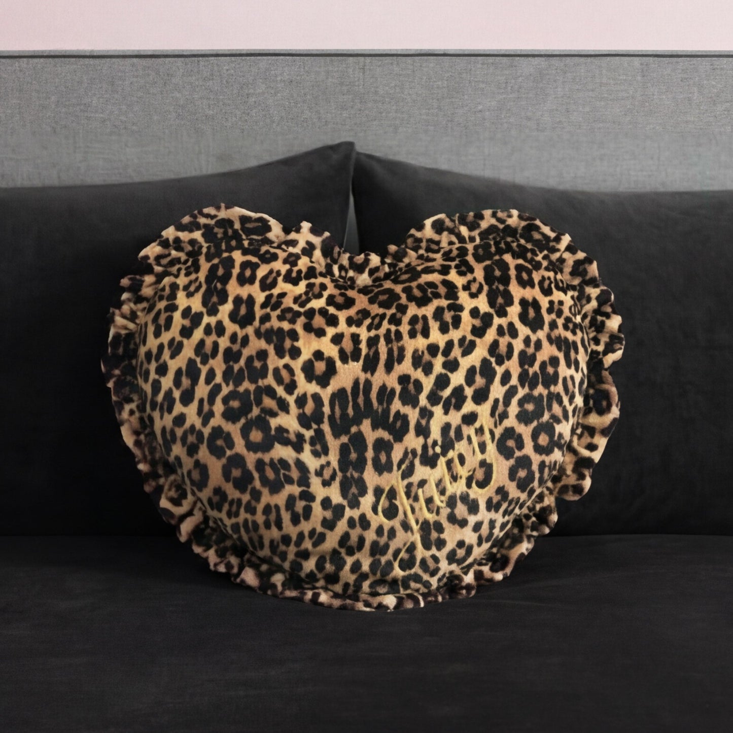 The Heart Shaped Decorative Pillow, featuring a ruffled edge and cheetah print inspired by Juicy Couture, rests on a dark gray velvet sofa with a gray backrest.