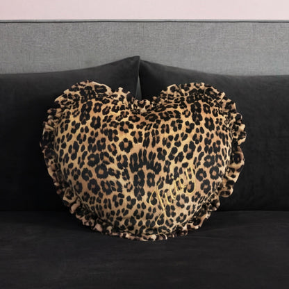 The Heart Shaped Decorative Pillow, featuring a ruffled edge and cheetah print inspired by Juicy Couture, rests on a dark gray velvet sofa with a gray backrest.