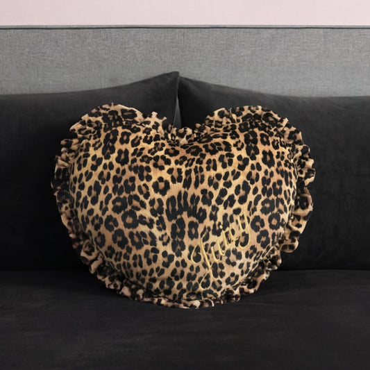 The Heart Shaped Decorative Pillow, featuring a ruffled edge and cheetah print inspired by Juicy Couture, rests on a dark gray velvet sofa with a gray backrest.