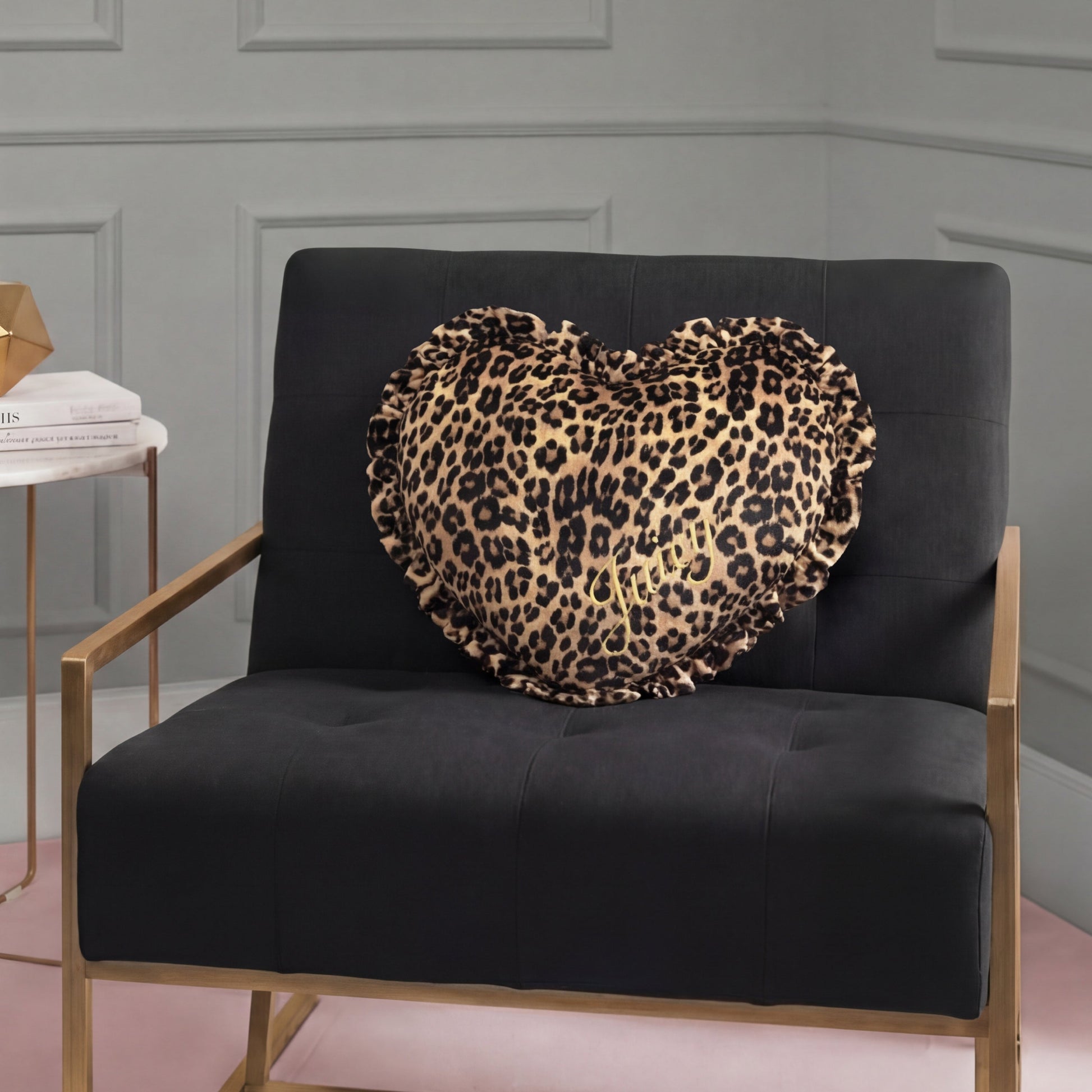 A black chair with gold arms holds a Heart Shaped Decorative Pillow with cheetah print and ruffled edges. The chair is on a pink rug beside a side table stacked with books in a gray-paneled room.
