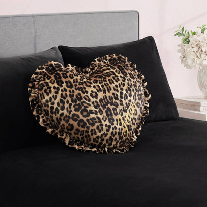 The Heart Shaped Decorative Pillow with a ruffled edge and cheetah print rests on a black bed with a gray headboard. In the background, there's a nightstand with white flowers in a vase and books.