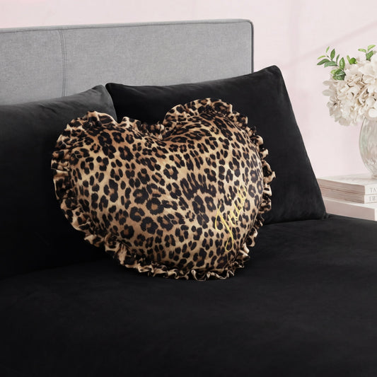 The Heart Shaped Decorative Pillow with a ruffled edge and cheetah print rests on a black bed with a gray headboard. In the background, there's a nightstand with white flowers in a vase and books.
