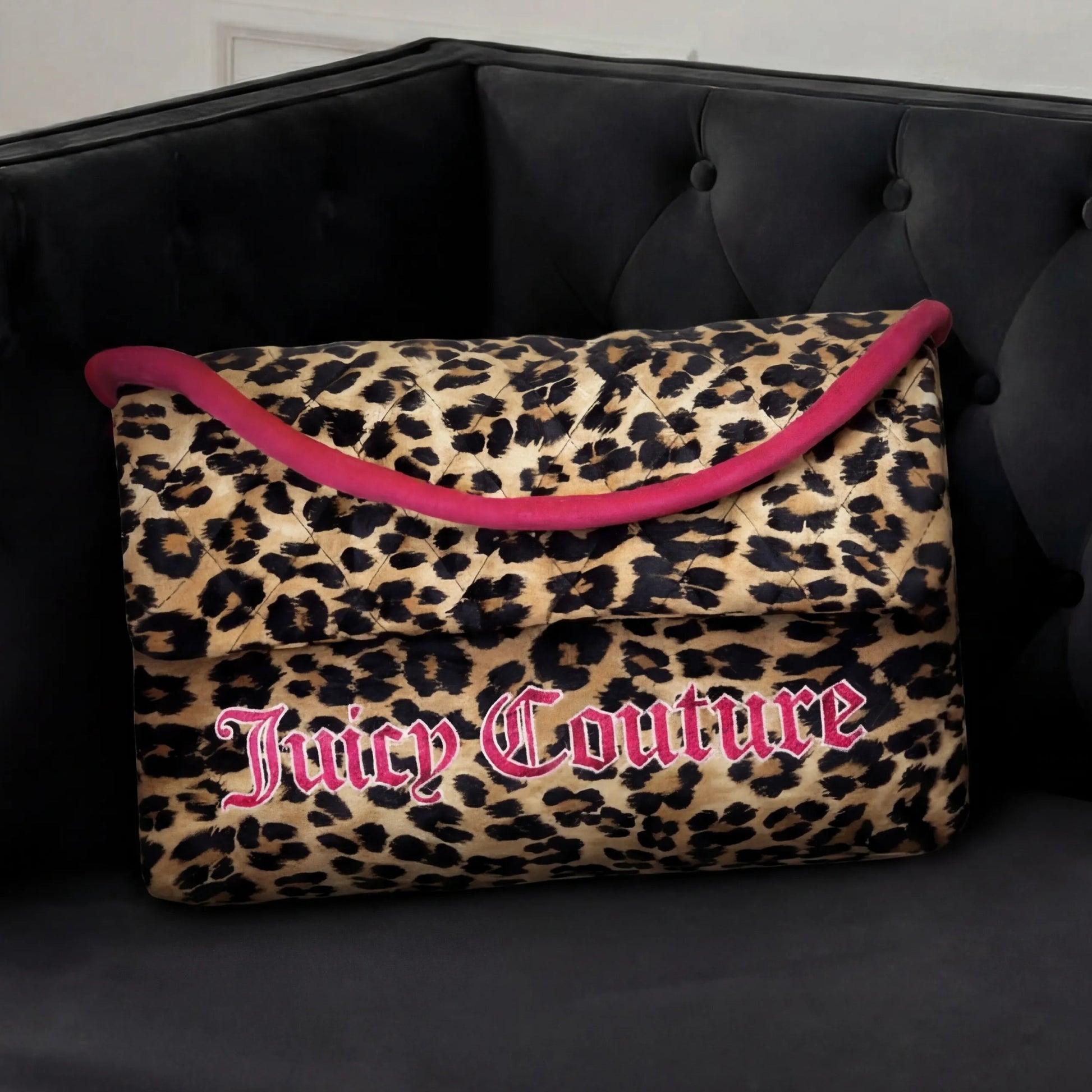 A Purse Decorative Pillow with leopard print, bold pink trim, and large Juicy Couture lettering sits on a tufted black velvet sofa, adding a trendy accent to any chic room.