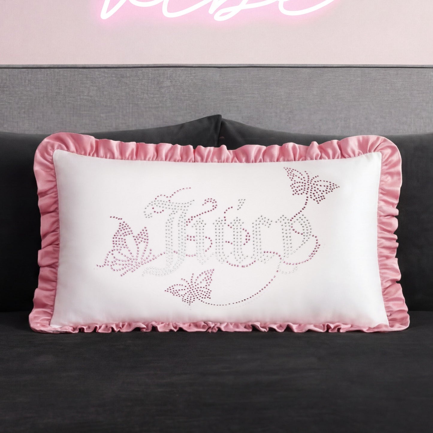 The Ruffle Decorative Pillow features pink ruffled edges, butterfly designs, and "fairy" in a decorative font on a pink and gray background with rhinestone accents, shown here on a dark sofa.