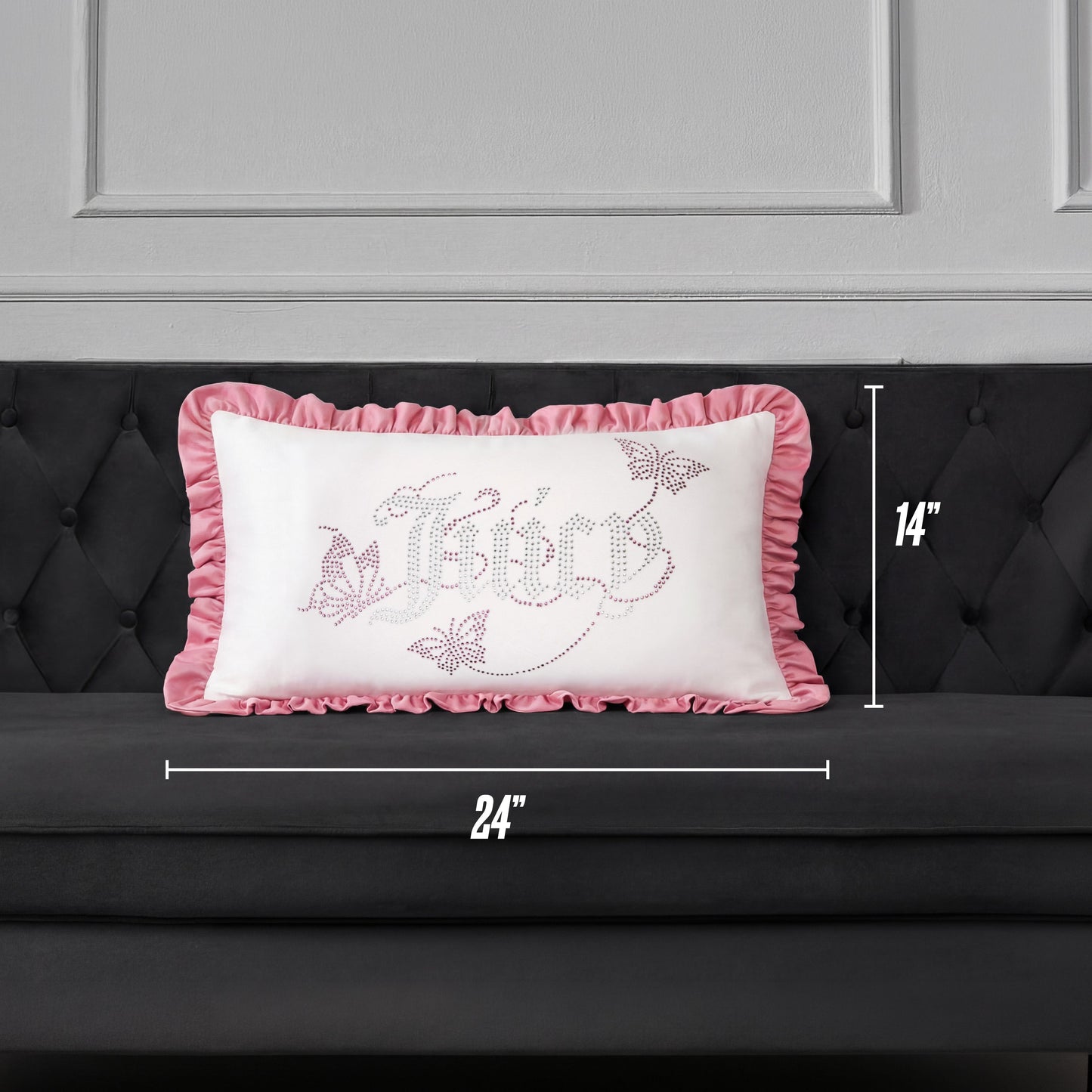A Ruffle Decorative Pillow by Juicy Couture, featuring pink ruffled edges and a decorative design, is placed on a dark gray couch. This rectangular pillow measures 24 inches wide by 14 inches tall.