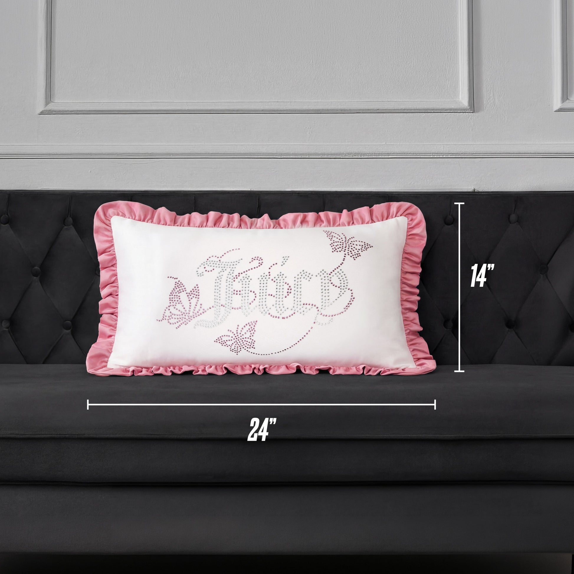 A Ruffle Decorative Pillow by Juicy Couture, featuring pink ruffled edges and a decorative design, is placed on a dark gray couch. This rectangular pillow measures 24 inches wide by 14 inches tall.