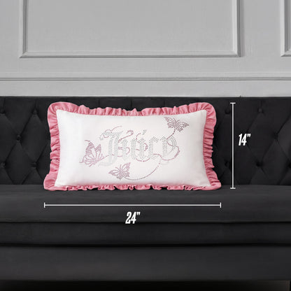 A Ruffle Decorative Pillow by Juicy Couture, featuring pink ruffled edges and a decorative design, is placed on a dark gray couch. This rectangular pillow measures 24 inches wide by 14 inches tall.