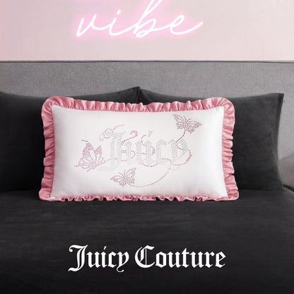 The Ruffle Decorative Pillow features white fabric, pink ruffled edges, and sparkling butterfly rhinestones, resting on a dark bed beneath a pink neon sign with Juicy Couture elegantly scripted at the bottom.
