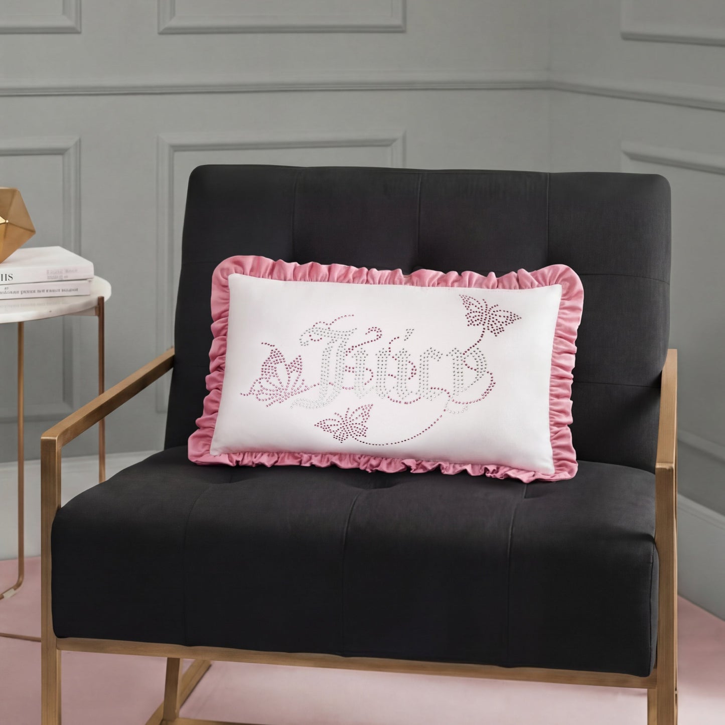A black chair with gold armrests holds a white Ruffle Decorative Pillow featuring pink ruffles, "Believe" text, butterflies, and floral designs in a modern room with gray paneled walls.